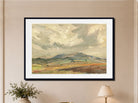 A framed landscape painting of rolling green and brown hills under a cloudy sky, displayed on a beige wall above a table with a plant and a lamp.