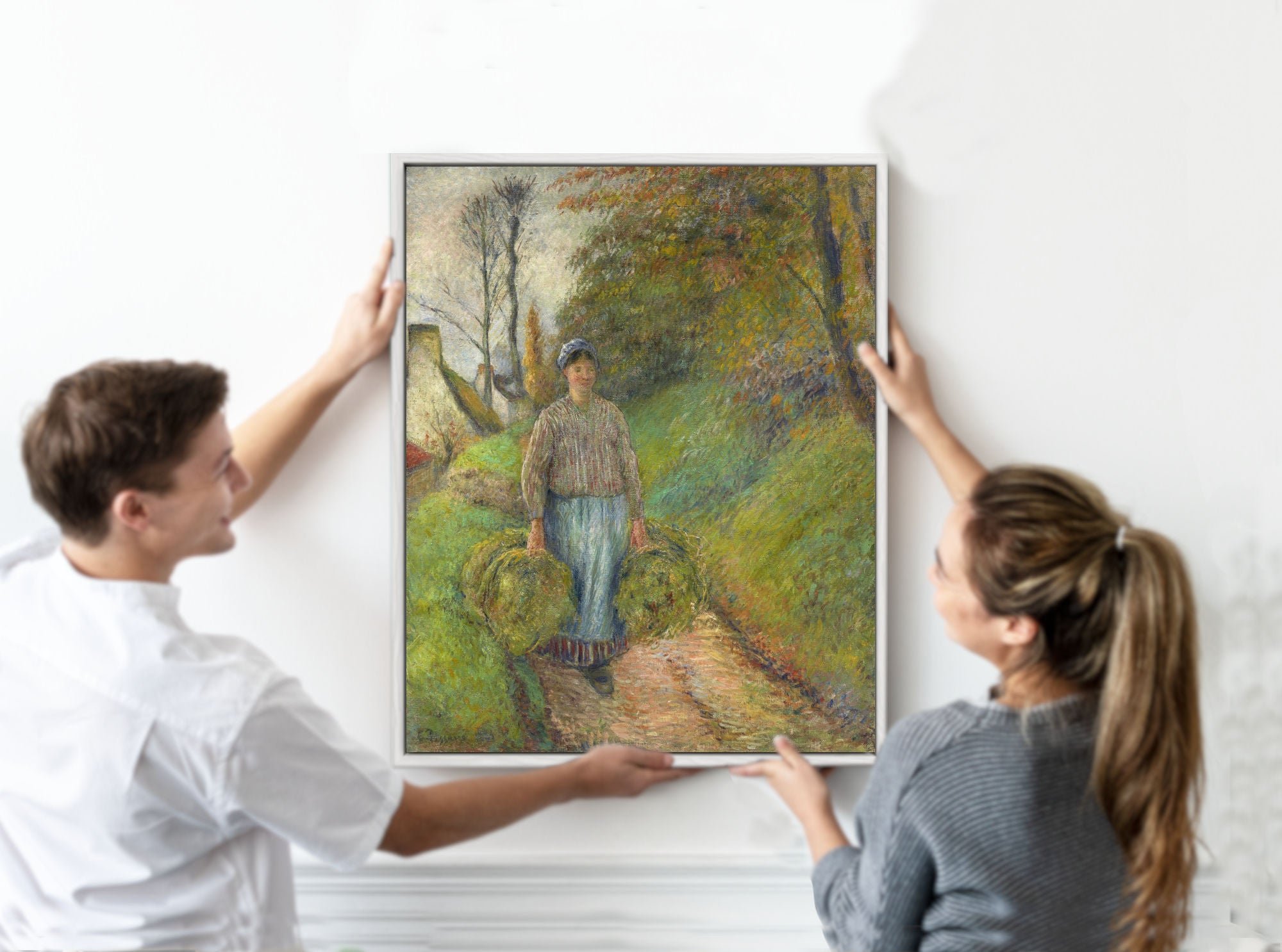 Two people hanging a framed painting of a person walking in a forest on a white wall.