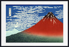 A framed Japanese-style art print depicting Mount Fuji, with a red mountain, blue sky, white clouds, and dark green foreground, shown in a black frame with a white mat.