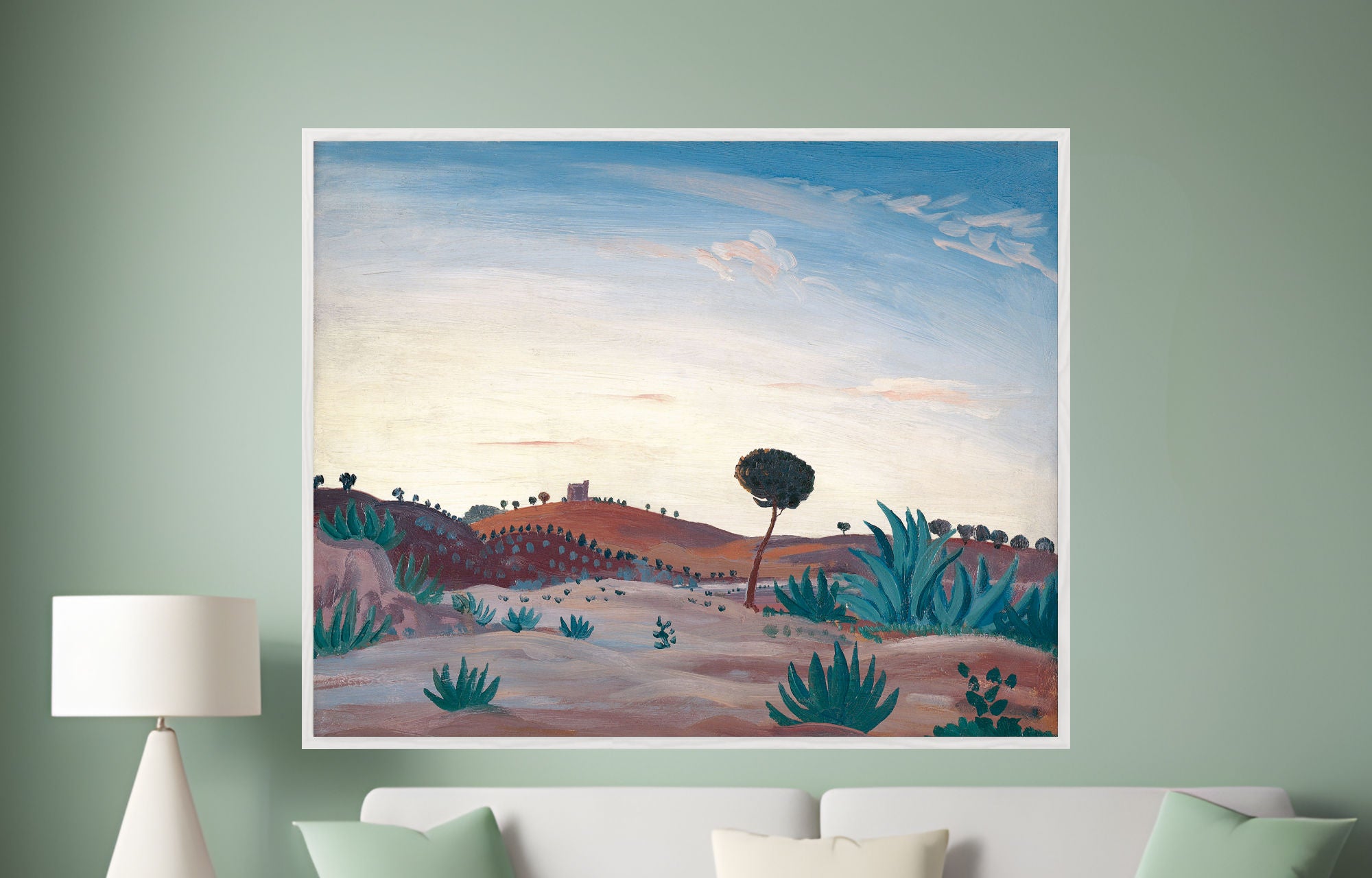 A framed landscape art print hanging on a sage green wall above a white sofa with mint green pillows. The painting depicts a desert scene with rolling hills, a lone tree, and blue-green cacti under a blue and cream sky.