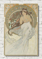 An Art Nouveau style poster print featuring a woman in a flowing white gown, mounted on a white brick wall. The artwork has ornate circular decorative elements and a warm cream, gold, and muted green palette.