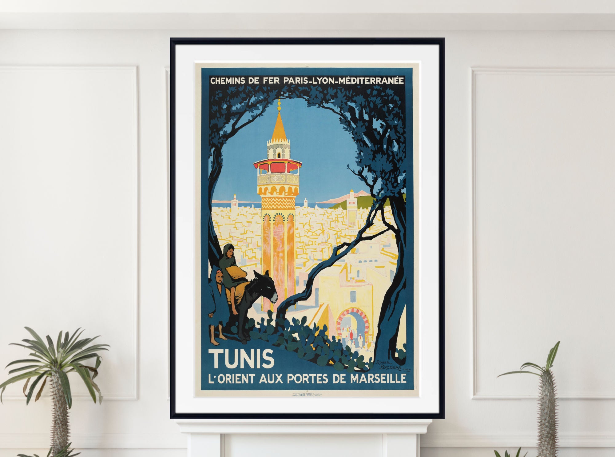 A framed vintage-style travel poster for Tunis, displayed on a white wall above a white mantel with small potted plants. The poster features a blue sky, a golden tower, and dark tree silhouettes, with French text including 'CHEMINS DE FER PARIS-LYON-MEDITERRANEEZ' and 'TUNIS L'ORIENT AUX PORTES DE MARSEILLE'.