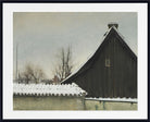 A framed art print depicting a winter landscape with a dark wooden barn, snow-covered roofs, bare trees, and a pale gray sky, shown with a wide white mat and black frame.