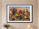 A framed still life painting of fruits, flowers, and a terracotta pot against a mountainous background, displayed on a beige wall above a small plant and lamp.