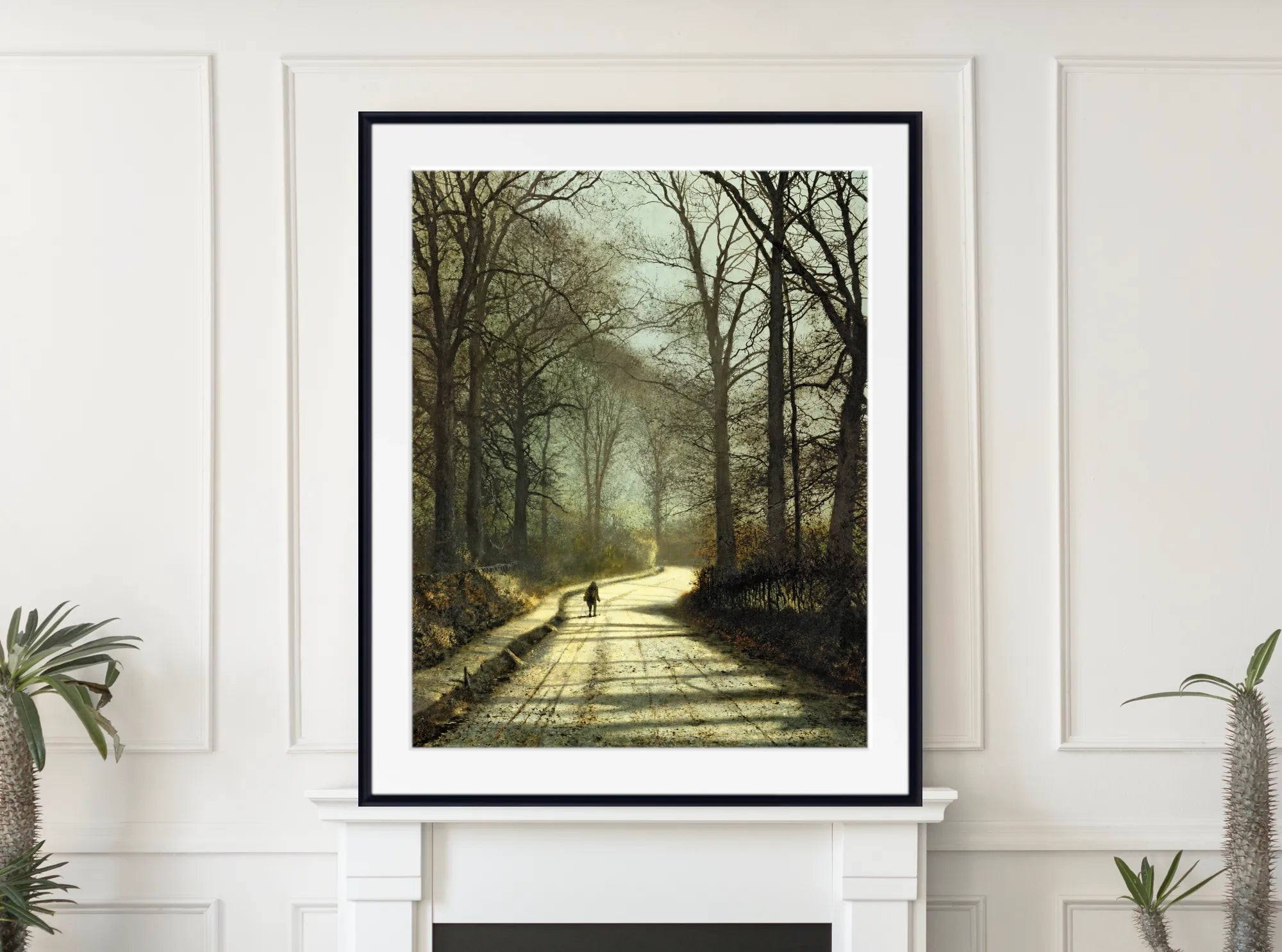 A framed art print depicting a misty forest path with bare trees and a distant cyclist, displayed on a white mantelpiece in a bright room.