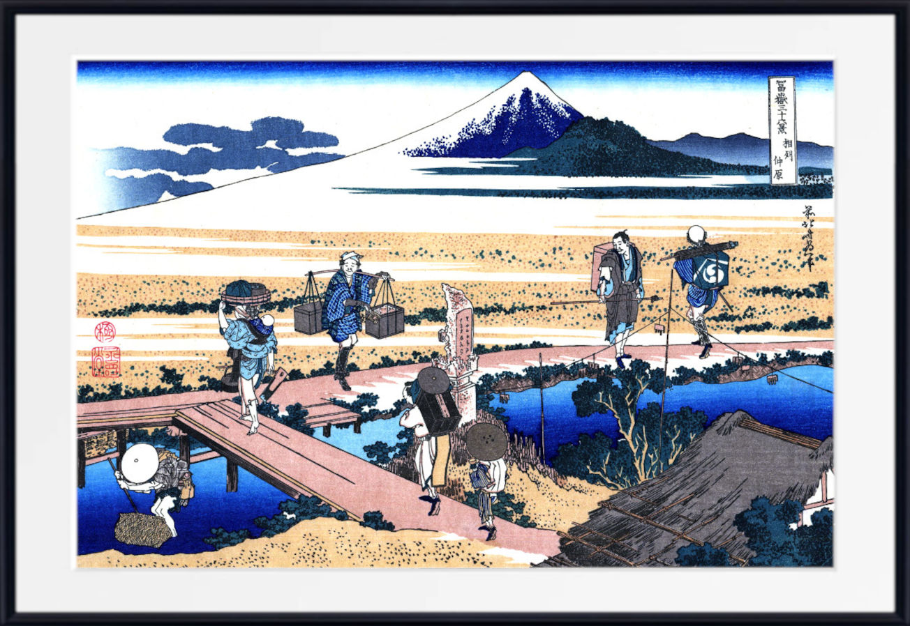 A framed Japanese ukiyo-e woodblock print depicting Mount Fuji, with figures walking across wooden bridges over blue water in the foreground. The print is shown with a wide white mat and a black frame.