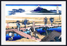 A framed Japanese ukiyo-e woodblock print depicting Mount Fuji, with figures walking across wooden bridges over blue water in the foreground. The print is shown with a wide white mat and a black frame.