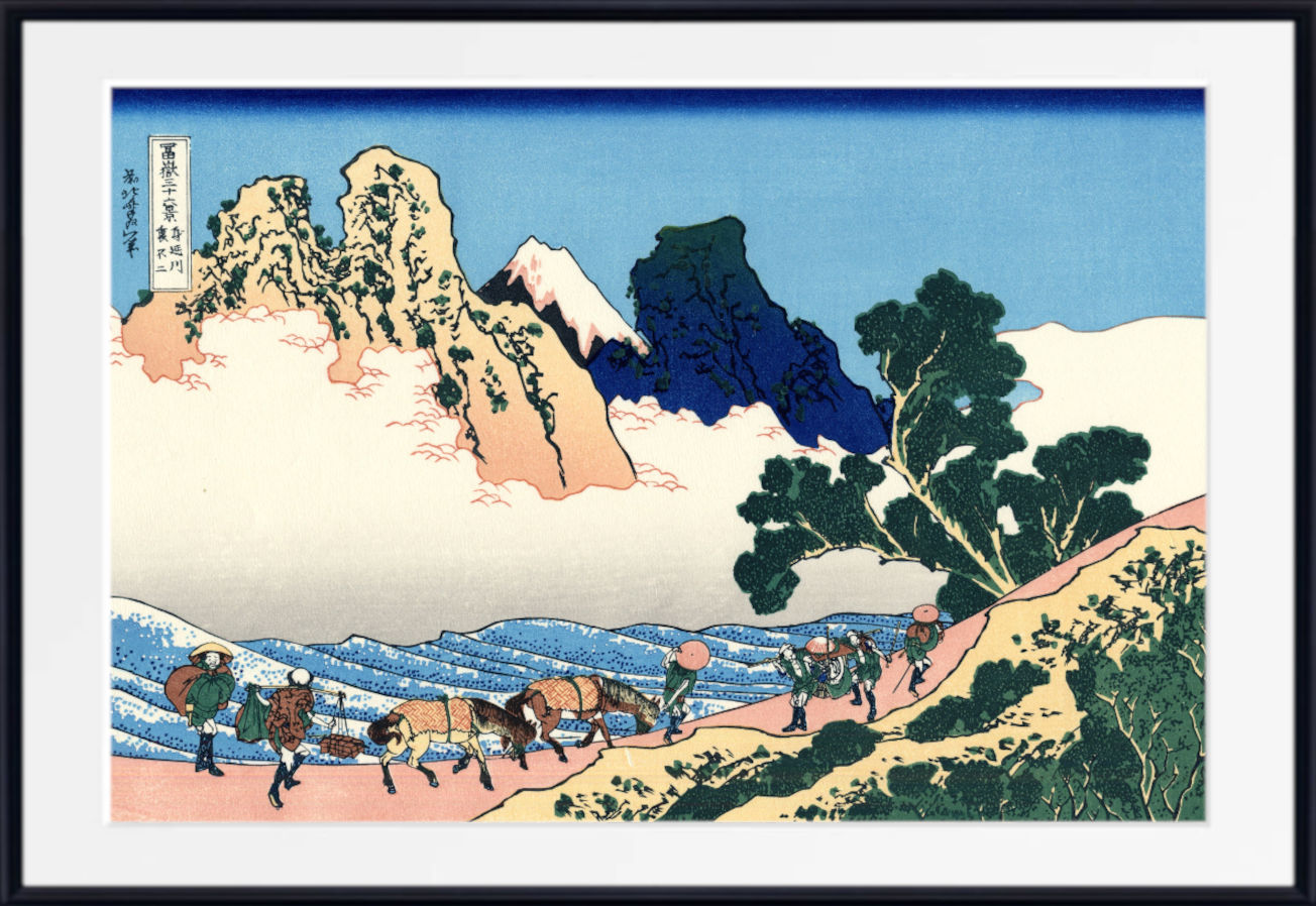 A framed Japanese-style art print showing a mountain landscape with a snowy peak, blue sky, waves, and a group of people and horses along a path. The print is presented with a white mat and a black frame.