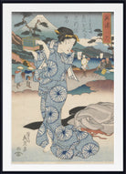 A framed Japanese ukiyo-e woodblock print featuring a woman in a blue patterned kimono standing in front of a coastal landscape with Mount Fuji, figures, boats, and buildings. The print is shown with a white mat and black frame.
